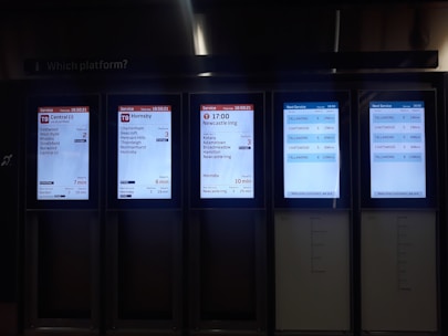 A set of digital train information screens displaying arrival and departure times, platforms, and destinations. Each screen provides different timetable details for various train routes, showing times in a clear digital format. The background is dimly lit, emphasizing the bright screens.