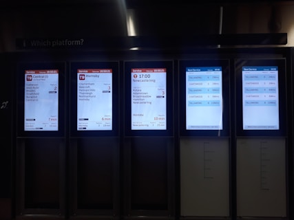 A set of digital train information screens displaying arrival and departure times, platforms, and destinations. Each screen provides different timetable details for various train routes, showing times in a clear digital format. The background is dimly lit, emphasizing the bright screens.