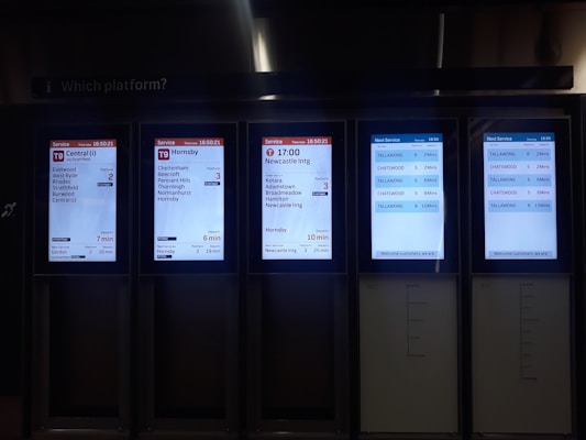 A set of digital train information screens displaying arrival and departure times, platforms, and destinations. Each screen provides different timetable details for various train routes, showing times in a clear digital format. The background is dimly lit, emphasizing the bright screens.