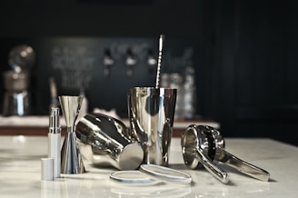 A collection of shiny metallic bar tools is arranged on a counter, including a cocktail shaker, jigger, and citrus press. The smooth, reflective surfaces create a sleek and modern appearance. The background is subtly blurred, with dark-colored elements that create depth.