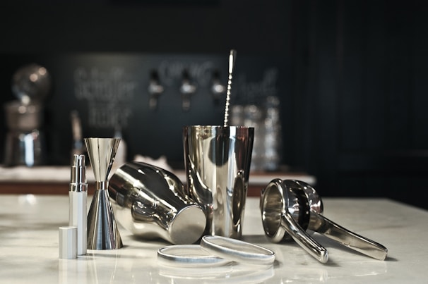 A collection of shiny metallic bar tools is arranged on a counter, including a cocktail shaker, jigger, and citrus press. The smooth, reflective surfaces create a sleek and modern appearance. The background is subtly blurred, with dark-colored elements that create depth.