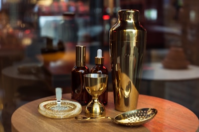 A collection of bar tools is neatly arranged on a wooden surface. The display includes a shiny golden cocktail shaker, a strainer, a jigger, and some brown glass bottles with droppers. The background is softly blurred, giving a warm, atmospheric feel.