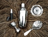 A collection of shiny silver bar tools including a cocktail shaker, strainer, jigger, and other accessories are arranged on a textured brown background resembling dried grass. The metallic surfaces reflect light, creating a polished appearance.