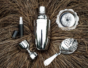 A collection of shiny silver bar tools including a cocktail shaker, strainer, jigger, and other accessories are arranged on a textured brown background resembling dried grass. The metallic surfaces reflect light, creating a polished appearance.