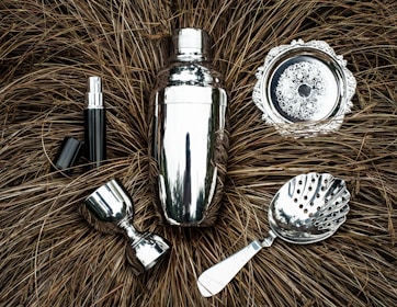 A collection of shiny silver bar tools including a cocktail shaker, strainer, jigger, and other accessories are arranged on a textured brown background resembling dried grass. The metallic surfaces reflect light, creating a polished appearance.