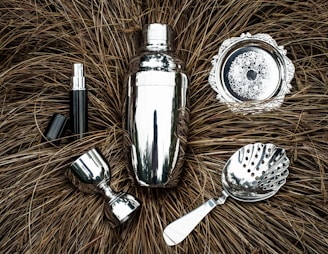 A collection of shiny silver bar tools including a cocktail shaker, strainer, jigger, and other accessories are arranged on a textured brown background resembling dried grass. The metallic surfaces reflect light, creating a polished appearance.
