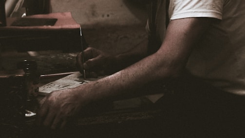 A person wearing a white shirt is engaged in a task involving a piece of paper and a pen or similar writing tool. The setting appears dimly lit, suggesting a focused or intimate atmosphere. The arm resting on a surface adds to the sense of concentration.