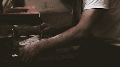 A person wearing a white shirt is engaged in a task involving a piece of paper and a pen or similar writing tool. The setting appears dimly lit, suggesting a focused or intimate atmosphere. The arm resting on a surface adds to the sense of concentration.