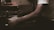 A person wearing a white shirt is engaged in a task involving a piece of paper and a pen or similar writing tool. The setting appears dimly lit, suggesting a focused or intimate atmosphere. The arm resting on a surface adds to the sense of concentration.