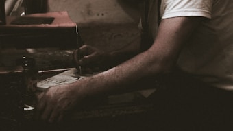 A person wearing a white shirt is engaged in a task involving a piece of paper and a pen or similar writing tool. The setting appears dimly lit, suggesting a focused or intimate atmosphere. The arm resting on a surface adds to the sense of concentration.