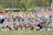 A large group of young women, all dressed in matching gray shirts and navy blue shorts, posing together outdoors. They are smiling and appear to be part of a sports team. The background shows a sports field with spectators in the stands.