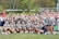 A large group of young women, all dressed in matching gray shirts and navy blue shorts, posing together outdoors. They are smiling and appear to be part of a sports team. The background shows a sports field with spectators in the stands.