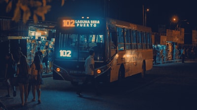 A city bus with the sign '102 Laguna Seca Puerto' is illuminated by its lights at night. People are walking nearby, and small shops with bright lights are visible in the background, indicating a busy area.