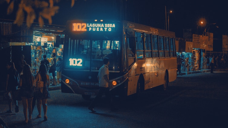 A city bus with the sign '102 Laguna Seca Puerto' is illuminated by its lights at night. People are walking nearby, and small shops with bright lights are visible in the background, indicating a busy area.