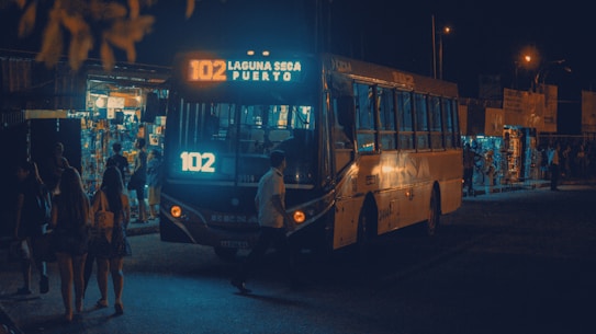 A city bus with the sign '102 Laguna Seca Puerto' is illuminated by its lights at night. People are walking nearby, and small shops with bright lights are visible in the background, indicating a busy area.