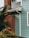 A section of a house with light blue wooden siding and a gray shingle roof. A red brick chimney is partially visible, surrounded by lush green bushes. A white gutter runs down the side of the house.