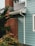A section of a house with light blue wooden siding and a gray shingle roof. A red brick chimney is partially visible, surrounded by lush green bushes. A white gutter runs down the side of the house.