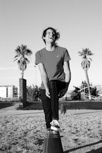 A person is balancing on one foot atop a cone structure while laughing heartily. The setting includes palm trees and a distant building, suggesting an outdoor location. The image is in black and white, highlighting contrasts between light and shadow.