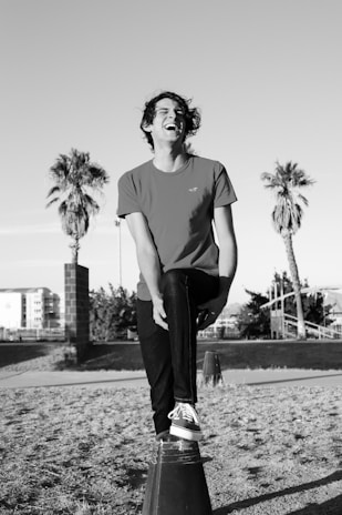 A person is balancing on one foot atop a cone structure while laughing heartily. The setting includes palm trees and a distant building, suggesting an outdoor location. The image is in black and white, highlighting contrasts between light and shadow.