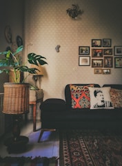 A cozy living room with a dark sofa adorned with decorative pillows, one featuring a monochrome portrait design. The room has a warm ambiance with various decorations on the wall, including framed photographs and art pieces. A patterned area rug covers part of the floor, and there's a wicker basket-style lamp near a potted plant with large green leaves. A small, classic wooden table supports the lamp.