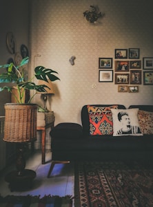 A cozy living room with a dark sofa adorned with decorative pillows, one featuring a monochrome portrait design. The room has a warm ambiance with various decorations on the wall, including framed photographs and art pieces. A patterned area rug covers part of the floor, and there's a wicker basket-style lamp near a potted plant with large green leaves. A small, classic wooden table supports the lamp.