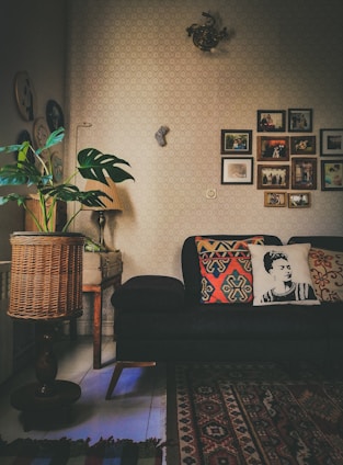 A cozy living room with a dark sofa adorned with decorative pillows, one featuring a monochrome portrait design. The room has a warm ambiance with various decorations on the wall, including framed photographs and art pieces. A patterned area rug covers part of the floor, and there's a wicker basket-style lamp near a potted plant with large green leaves. A small, classic wooden table supports the lamp.