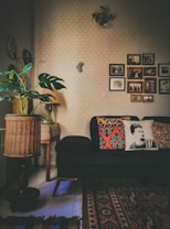 A cozy living room with a dark sofa adorned with decorative pillows, one featuring a monochrome portrait design. The room has a warm ambiance with various decorations on the wall, including framed photographs and art pieces. A patterned area rug covers part of the floor, and there's a wicker basket-style lamp near a potted plant with large green leaves. A small, classic wooden table supports the lamp.
