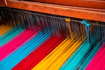 Colorful threads are arranged on a loom, showcasing vibrant hues of blue, red, yellow, and pink. The threads are tightly lined up and prepared for weaving, with wooden parts of the loom visible at the top.