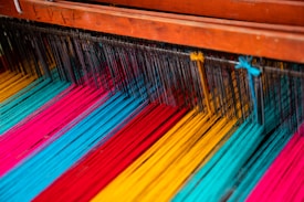 Colorful threads are arranged on a loom, showcasing vibrant hues of blue, red, yellow, and pink. The threads are tightly lined up and prepared for weaving, with wooden parts of the loom visible at the top.