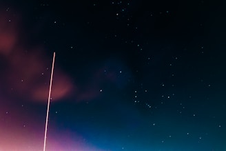 A night sky filled with stars, featuring a prominent vertical light trail from the bottom left corner extending towards the sky. The combination of deep black, blue, and hints of reddish clouds creates a serene celestial scene.