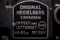 A metal plaque with the text 'Original Heidelberg Einfarben Offset oder Letterset' and measurements '46x62 cm' and '18x24½'. The background of the plaque is dark, and the text and borders are in a lighter metallic color.