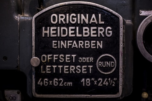 A metal plaque with the text 'Original Heidelberg Einfarben Offset oder Letterset' and measurements '46x62 cm' and '18x24½'. The background of the plaque is dark, and the text and borders are in a lighter metallic color.