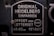 A metal plaque with the text 'Original Heidelberg Einfarben Offset oder Letterset' and measurements '46x62 cm' and '18x24½'. The background of the plaque is dark, and the text and borders are in a lighter metallic color.
