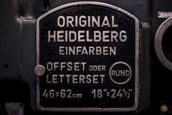 A metal plaque with the text 'Original Heidelberg Einfarben Offset oder Letterset' and measurements '46x62 cm' and '18x24&frac12;'. The background of the plaque is dark, and the text and borders are in a lighter metallic color.