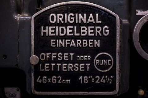 A metal plaque with the text 'Original Heidelberg Einfarben Offset oder Letterset' and measurements '46x62 cm' and '18x24½'. The background of the plaque is dark, and the text and borders are in a lighter metallic color.