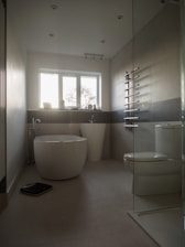 A modern bathroom with a minimalist design, featuring a freestanding bathtub, a large white pedestal sink, and a toilet. The room has light beige tiles and a glass-walled shower area. A towel radiator is mounted on the wall, and a digital scale is placed on the floor. The window allows natural light to enter, illuminating the space.