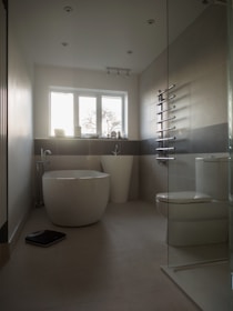 A modern bathroom with a minimalist design, featuring a freestanding bathtub, a large white pedestal sink, and a toilet. The room has light beige tiles and a glass-walled shower area. A towel radiator is mounted on the wall, and a digital scale is placed on the floor. The window allows natural light to enter, illuminating the space.