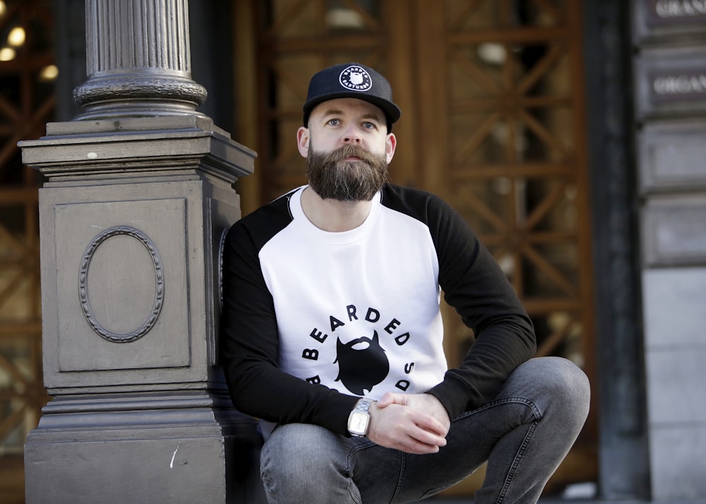 man in white and black raglan shirt and gray pants leaning on black column