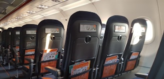 Rows of airplane seats are visible, with tray tables and safety instruction cards visible in the pocket of each seat. The interior is well-lit, with natural light coming through the windows on one side.