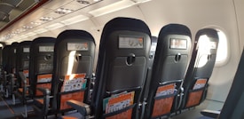 Rows of airplane seats are visible, with tray tables and safety instruction cards visible in the pocket of each seat. The interior is well-lit, with natural light coming through the windows on one side.