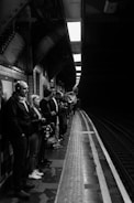 A group of people stand on a subway platform, waiting for the train. The setting is a dimly lit underground station, with patterned tiles and structural beams visible above. The platform edge is marked, indicating where passengers should stand back. The atmosphere appears calm as individuals engage with their phones or stare into the distance.