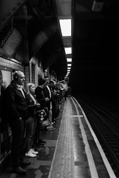A group of people stand on a subway platform, waiting for the train. The setting is a dimly lit underground station, with patterned tiles and structural beams visible above. The platform edge is marked, indicating where passengers should stand back. The atmosphere appears calm as individuals engage with their phones or stare into the distance.
