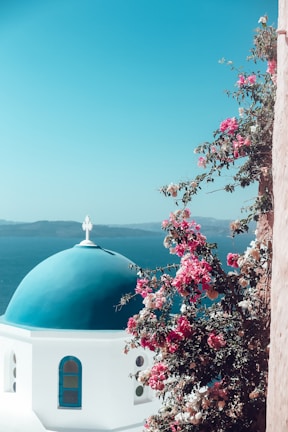 A picturesque view of a bright blue domed building characteristic of traditional architecture in the Greek islands, with vibrant pink and white bougainvillea flowers climbing up the wall. The background features a deep blue sea and distant land.