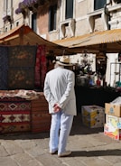 A person wearing a white suit and straw hat stands with hands behind their back, observing a market stall. Colorful textiles and patterned rugs are displayed, and the background features a historic stone building with decorative windows adorned with flowering plants. Another market stall under a striped canopy is visible, containing various glass items and boxes.