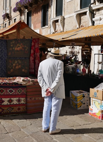 A person wearing a white suit and straw hat stands with hands behind their back, observing a market stall. Colorful textiles and patterned rugs are displayed, and the background features a historic stone building with decorative windows adorned with flowering plants. Another market stall under a striped canopy is visible, containing various glass items and boxes.