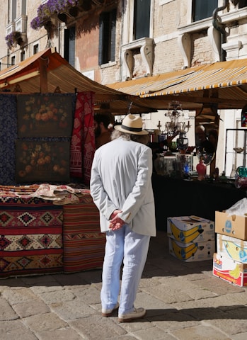 A person wearing a white suit and straw hat stands with hands behind their back, observing a market stall. Colorful textiles and patterned rugs are displayed, and the background features a historic stone building with decorative windows adorned with flowering plants. Another market stall under a striped canopy is visible, containing various glass items and boxes.