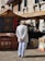 A person wearing a white suit and straw hat stands with hands behind their back, observing a market stall. Colorful textiles and patterned rugs are displayed, and the background features a historic stone building with decorative windows adorned with flowering plants. Another market stall under a striped canopy is visible, containing various glass items and boxes.