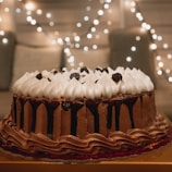 A beautifully decorated chocolate cake with rich layers of chocolate frosting and a topping of whipped cream peaks and dark cherries. The background features soft, warm string lights creating a cozy and festive atmosphere.