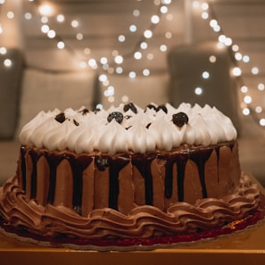 A beautifully decorated chocolate cake with rich layers of chocolate frosting and a topping of whipped cream peaks and dark cherries. The background features soft, warm string lights creating a cozy and festive atmosphere.