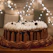 A smiling woman holding a beautifully decorated chocolate ganache cake in a cozy kitchen setting.